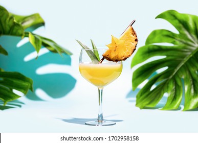 Summer refreshing alcoholic cocktail concept, front view of a garnished ready-to-drink pineapple margarita glass in front of palm leaves in a shining sun