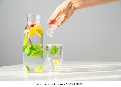 Summer Refreshing Drink. Water With Lemon, Lime And Mint. Woman Hand Drops Ice Cubes Into Glass With Alcohol Free Beverage. Studio Shot.