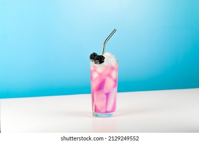 Summer refreshing pink cocktail with ice cube, metal straw and blackberry on blue background.