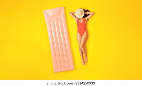 Summer Relax. Woman In One-Piece Swimsuit Posing Lying Near Inflatable Lilo Mattress, Covering Face From Sun With Hat, Relaxing And Sunbathing In Studio Over Yellow Background, Top View. Panorama