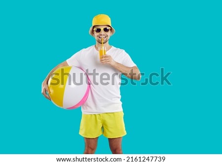 Summer rest. Happy male beach vacationer enjoys freshly squeezed orange juice isolated on light blue background. Smiling man in summer clothes and with inflatable beach ball drinks juice through straw