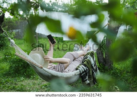 Summer Retreat: Lady in a Sun Hat Enjoys Her Green Oasis, Leisurely Scrolling Through Social Media on Her Smartphone