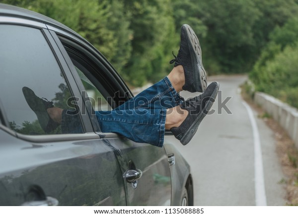 Summer road trip car vacation\
concept. Woman legs out the windows in car above the road in\
forest. Conceptual freedom, travel and holidays image with copy\
space.