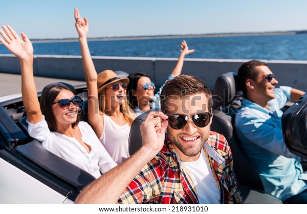 Summer road trip. Group of young happy people\
enjoying road trip in their convertible while handsome man\
adjusting his sunglasses and\
smiling