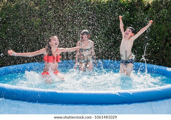 Summer season concept background.  Three happy\
school children friends - one boy, two girls - have fun making\
water splashes in inflatable garden swimming pool, refreshing\
themselves in hot\
weather