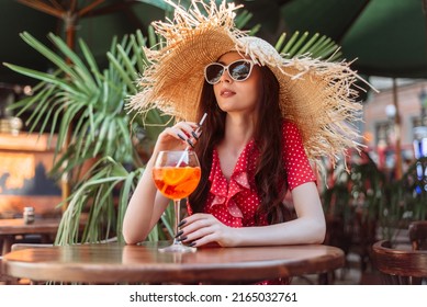 Summer season. Cute girl drink cold cocktail aperol spritz on the terrace. Summertime lifestyle portrait of young pretty woman in sunglasses, straw hat and red polka-dot