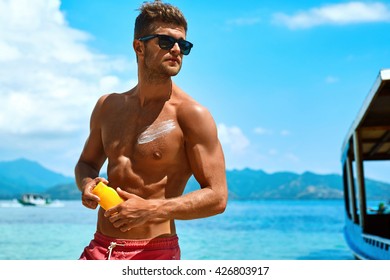 Summer Skin Care. Handsome Sexy Man In Fashionable Sunglasses Tanning Using Protective Sunscreen Lotion On Athletic Muscular Body. Male Model Sunbathing With Solar Sun Block Protection Cream On Beach