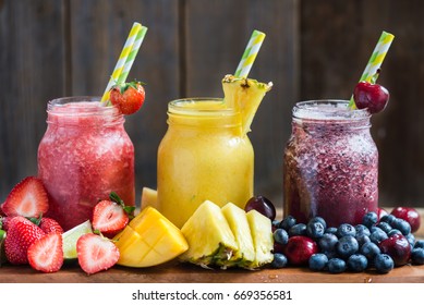 Summer Slushies from Blueberries, Cherries, Lemon, Mango, Strawberries, Lime, Pineapple and Ice with Ingredients nearby, on dark rustic background