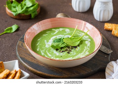 Summer spinach soup with cream in a pink plate on a wooden board on a brown concrete background. Spinach recipes. Healthy food