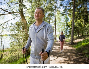 Summer Sport In Finland - Nordic Walking. Man And Mature Woman Hiking In Green Sunny Forest. Active People Outdoors. Scenic Peaceful Finnish Summer Landscape.