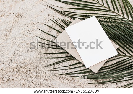 Summer stationery still life. Closeup of blank card mock-up and craft envelope on green date palm leaves. Sandy beach background or desert. Flat lay, top view. Tropical vacation concept. 