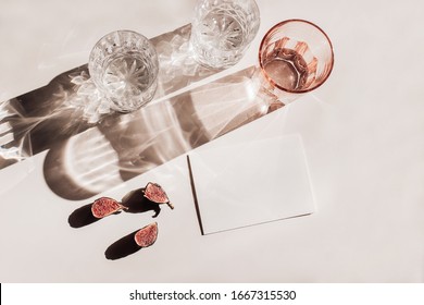 Summer stationery still life scene. Glasses of water, cut figs fruit. Pink table background in sunlight. Blank business, greeting card, invitation mockup scene. Long harsh shadows. Flat lay, top view