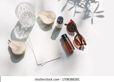 Summer stationery still life scene. Glass of water, cut pear fruit, cosmetic oil, cream bottles and sunglasses in sunlight. White table. Blank paper cards, invitations mockup scene, branches shadows.