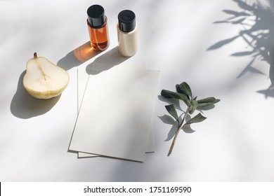 Summer Stationery Still Life Scene. Cut Pear Fruit, Cosmetic Oil, Cream Bottles And Olive Tree Branch. White Table. Blank Paper Cards, Invitations Mockup Scene. Long Shadows Overlay. Flat Lay, Top