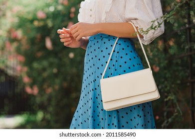Summer street fashion details: close up of  classic white faux leather bag, small handbag in elegant outfit. Woman wearing trendy polka dot blue midi skirt,  posing in street. Copy, empty  space