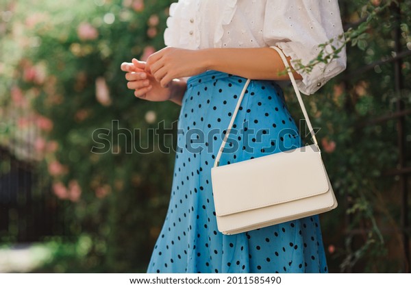 Summer street fashion details: close up of  classic\
white faux leather bag, small handbag in elegant outfit. Woman\
wearing trendy polka dot blue midi skirt,  posing in street. Copy,\
empty  space