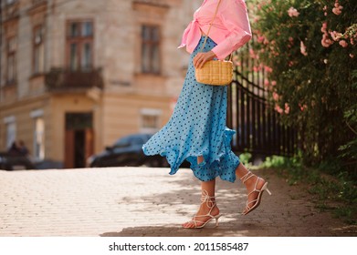 Summer street fashion details: elegant woman wearing trendy pink shirt with knot, polka dot blue midi skirt, white strap sandals, with yellow wicker leather shoulder bag, walking in street. Copy space
