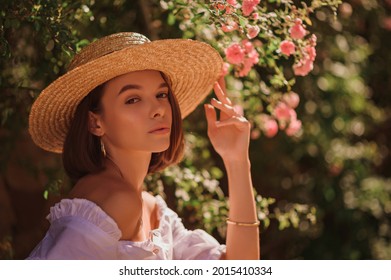 Summer street style, fashion portrait of beautiful brunette woman wearing stylish straw hat, white blouse, posing in street, near blooming roses. Copy, empty space for text
