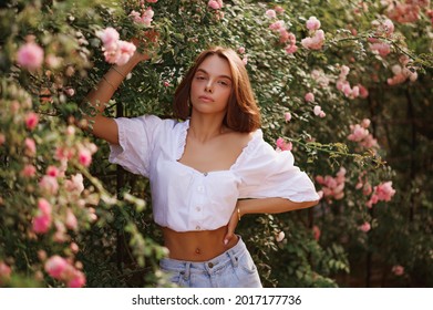 Summer street style, fashion portrait of beautiful lady wearing trendy summer outfit with  stylish  white blouse, denim shorts, posing in street, near blooming roses. Copy, empty space for text
