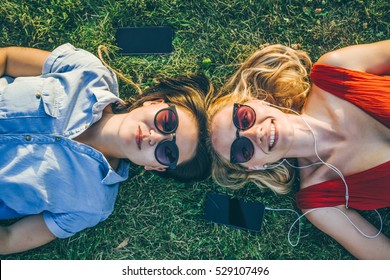 Summer sunny day.View from above.Two young smiling woman in sunglasses lying on green lawn heads together.Girl listens to music on headphones with your smartphone.Girls resting on grass in park.