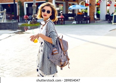 Summer sunny lifestyle fashion portrait of young stylish hipster woman walking on the street, wearing cute trendy outfit, drinking tasty smoothie, smiling enjoy her weekends, travel with backpack/