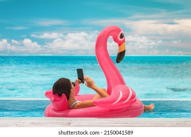 Summer swimming pool vacation relaxing woman floating in flamingo inflatable float using mobile phone at luxury resort sunbathing. Caribbean travel vacation hotel lifestyle