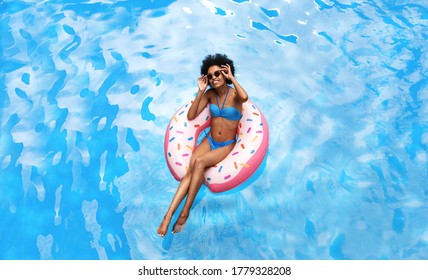 Summer time concept. Beautiful black woman in blue bikini swimming on inflatable ring at pool during tropical vacation, above view