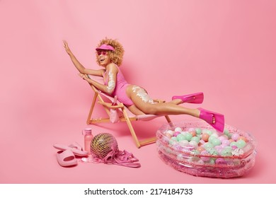 Summer Time Skin Care And Resort Concept. Cheerful Slim Curly Female Model Applies Sun Protection Lotion On Body Dressed In Bathingsuit Lies On Comfortable Deck Chair Spends Vacation At Seaside.