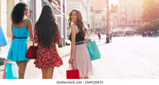 Summer In The Town. Three Young Beautiful Girls In Sophisticated Dresses Are Walking Along The City Streets After Shopping, One Girl Turns Around And Smiles Broadly.