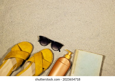 Summer And Travel Concept, Top View  Slippers, Body Cream And Sunglasses And Book On Sand