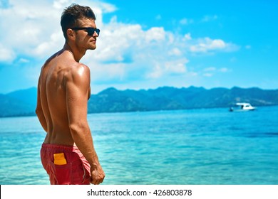 Summer Travel Vacation. Handsome Man With Sexy Body In Fashionable Sunglasses Sunbathing, Tanning At Sea Beach. Fitness Male Model With Sunscreen Lotion, Sun Block Skin Protection Cream In Pocket