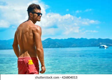 Summer Travel Vacation. Handsome Man With Sexy Body In Fashionable Sunglasses Sunbathing, Tanning At Sea Beach. Fitness Male Model With Sunscreen Lotion, Sun Block Skin Protection Cream In Pocket