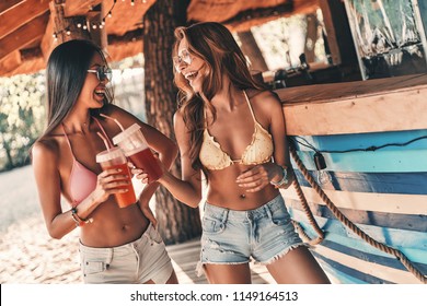 To the summer! Two attractive young women smiling and toasting each other while standing near the bar counter