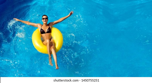 Summer vacation. Beautiful slim young woman enjoying an aqua park floating in a yellow big ring on sparkling blue pool while smiling at camera, panorama top view with copy space