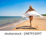 Summer vacation. Beautiful young woman in colorful swimsuit hat and sunglasses walking with a white scarf fluttering in the wind on the sandy beach on a beautiful sunny day. Back view 
