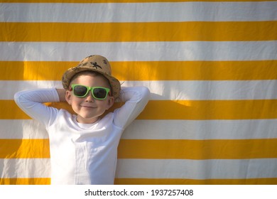Summer vacation. Boy in turquoise sunglasses lies on a orange towel against the background of the beach and the stove.