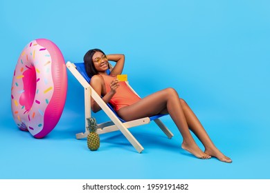 Summer vacation budget. Pretty black woman in suimsuit relaxing in lounge chair with credit card on blue studio background, full length. African American lady saving money for beach holiday