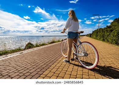 Summer vacation by bike. Young woman with long hair wearing white shirt and denim shorts standing with white and red fixed gear bike on bicycle path on seafront on sunny day.  - Powered by Shutterstock