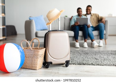 Summer Vacation Concept. African American Family Using Laptop While Packed Suitcase And Bags Lying On Floor At Home. Selective Focus On Luggage. Couple Booking Flight Tickets Online Via Computer