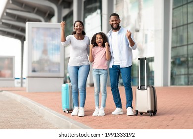 Summer Vacation Concept. Cheerful positive black family of three people having fun and shaking clenched fists, making winner gesture, travelling together standing near airport terminal with baggage - Shutterstock ID 1991333192