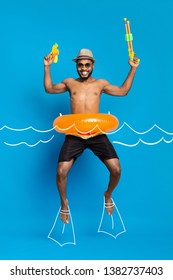 Summer Vacation Concept. Happy African American Guy In Swimwear And Inflatable Ring Jumping With Water Guns Like Floating In Water, Drawn Waves And Flippers