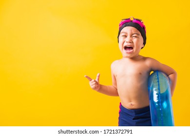 Summer Vacation Concept, Portrait Asian Happy Cute Little Child Boy Wear Goggles And Swimsuit Hold Blue Inflatable Ring, Kid Hav Fun Point Finger To Side Away, Studio Shot Isolated Yellow Background