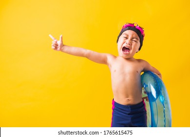 Summer Vacation Concept, Portrait Asian Happy Cute Little Child Boy Wear Goggles And Swimsuit Hold Blue Inflatable Ring, Kid Hav Fun Point Finger To Side Away, Studio Shot Isolated Yellow Background