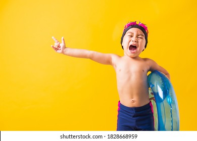 Summer Vacation Concept, Portrait Asian Happy Cute Little Child Boy Wear Goggles And Swimsuit Hold Blue Inflatable Ring, Kid Hav Fun Point Finger To Side Away, Studio Shot Isolated Yellow Background