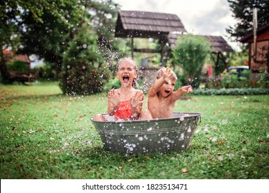 Summer vacation in countryside, local staycation. Cute kids having fun, bathing and splashing in old iron bowl on the backyard of wooden house, happy summertime, outdoor lifestyle, selective focus