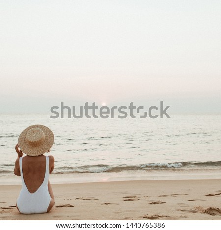 Summer vacation fashion concept. Young, tanned woman wearing a beautiful white swimsuit with a straw hat is sitting and relaxing on tropical beach with white sand and is watching sunset and sea.