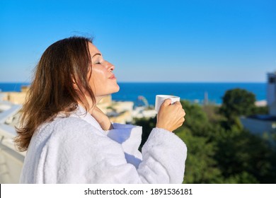 Summer vacation of happy mature woman. Middle-aged female in white bathrobe on balcony of hotel in seaside resort town. Enjoying cup of coffee, sun, beautiful seascape, copy space blue sky sea - Powered by Shutterstock