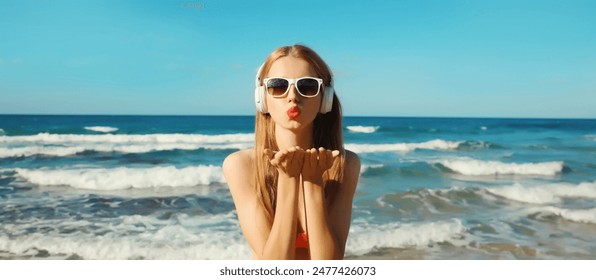 Summer vacation of happy young woman relaxing listening to music with headphones, modern girl blowing a kiss having fun on the beach at sea coast with waves on a sunny day - Powered by Shutterstock