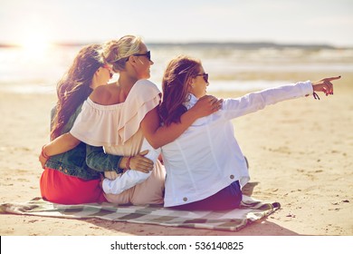 Summer Vacation, Holidays, Travel And People Concept - Group Of Smiling Young Women In Sunglasses Sitting On Beach Blanket And Pointing Finger To Something