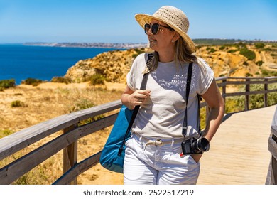 Summer vacation in Lagos Portugal. Young woman in beige casual clothes and sun hat carrying camera walking on wooden pier on cliffs during sightseeing Ponta da Piedade on beautiful sunny day. - Powered by Shutterstock